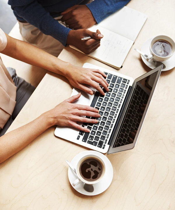 A top-down view of two people working together at a wooden table. One person types on a laptop, while the other writes in a notebook. Two cups of coffee sit nearby.